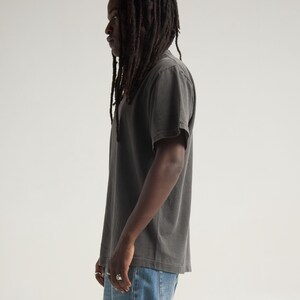 May include: A charcoal gray short-sleeved t-shirt with a crew neckline and a relaxed fit, paired with light blue denim jeans. The t-shirt features a solid, muted color tone and a standard hemline, worn with silver rings on the fingers.
