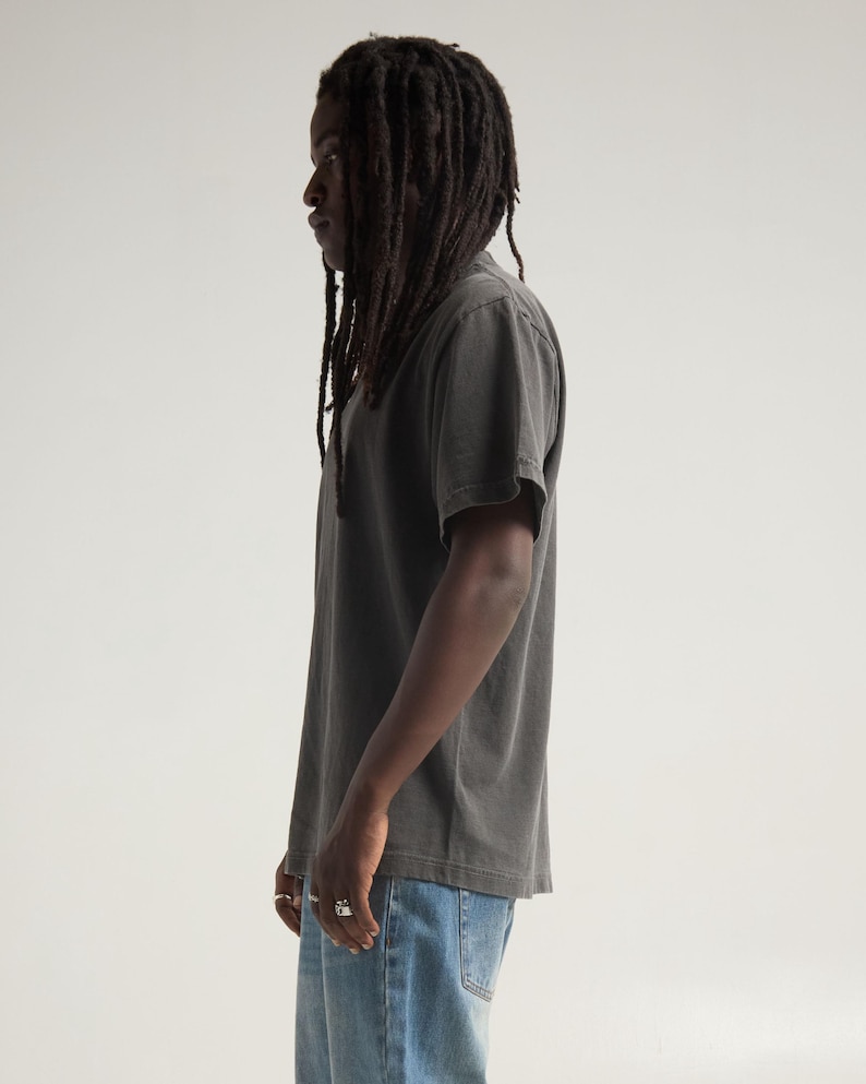 May include: A charcoal gray short-sleeved t-shirt with a crew neckline and a relaxed fit, paired with light blue denim jeans. The t-shirt features a solid, muted color tone and a standard hemline, worn with silver rings on the fingers.
