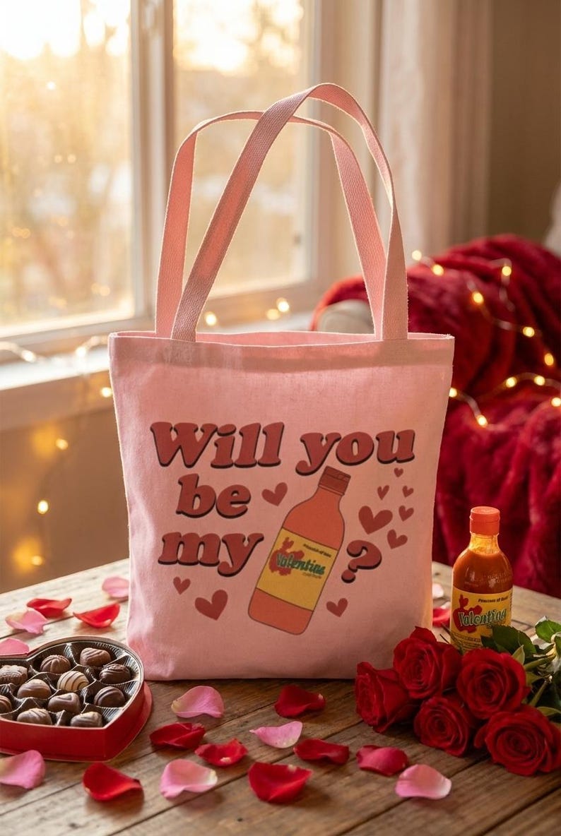 Op de afbeelding: Een roze draagtas met de tekst "Will you be my" en een afbeelding van een hete sausfles. De afbeelding bevat Valentijnsdag-items: rozen, chocolaatjes en bloemblaadjes.