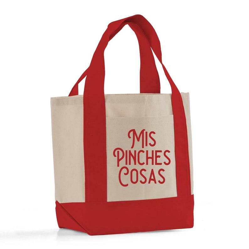 May include: A canvas tote bag with a cream-colored body and red accents on the bottom, handles, and pocket. The pocket features the phrase "Mis Pinches Cosas" in red script.