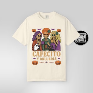 May include: Cream-colored t-shirt featuring a Halloween-themed graphic. The design includes three skeleton figures with colorful hair, holding cups and a ghost. The text "CAFECITO Y BRUJER&Iacute;A" is printed below the figures, with pumpkins and bats.