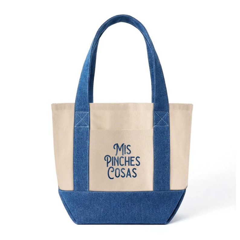 K&ouml;nnte beinhalten: Eine Canvas-Tragetasche mit jeansblauen Akzenten. Die Tasche hat einen cremefarbenen K&ouml;rper, jeansblaue Griffe und einen jeansblauen Boden. Der Text "Mis Pinches Cosas" ist blau auf der Vorderseite gestickt. Ein praktisches und stilvolles Accessoire.
