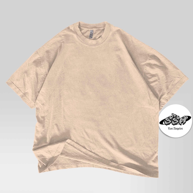 Pu&ograve; includere: Una maglietta beige oversize con scollo tondo. La maglietta &egrave; realizzata in un materiale morbido e ha maniche corte. Un logo circolare bianco con la scritta "SSA Los Angeles" si trova nell'angolo in basso a destra.
