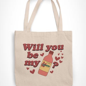 Op de afbeelding: Beige draagtas met de tekst "Will you be my" in rode letters met een bruine omlijning. Een fles Valentina-chilisaus en rode harten staan ook op de tas. De tas heeft twee beige handvatten.