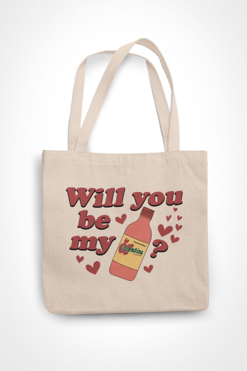 Op de afbeelding: Beige draagtas met de tekst "Will you be my" in rode letters met een bruine omlijning. Een fles Valentina-chilisaus en rode harten staan ook op de tas. De tas heeft twee beige handvatten.