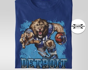 Op de afbeelding: Een blauw t-shirt met een afbeelding van een leeuw die een voetbaluniform draagt en een voetbal vasthoudt. De leeuw rent en er zijn bliksemschichten achter hem. De tekst "DETROIT" is onder de leeuw gedrukt.