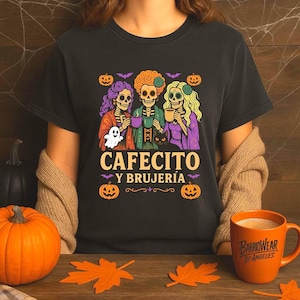 May include: Black t-shirt with a Halloween-themed graphic. The design features three skeleton figures holding mugs, with the text "CAFECITO Y BRUJER&Iacute;A" below. Pumpkins, bats, and a ghost complete the design. An orange mug is visible.