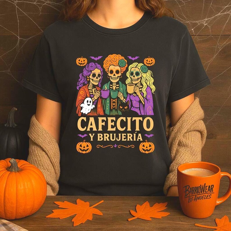 May include: Black t-shirt with a Halloween-themed graphic. The design features three skeleton figures holding mugs, with the text "CAFECITO Y BRUJER&Iacute;A" below. Pumpkins, bats, and a ghost complete the design. An orange mug is visible.