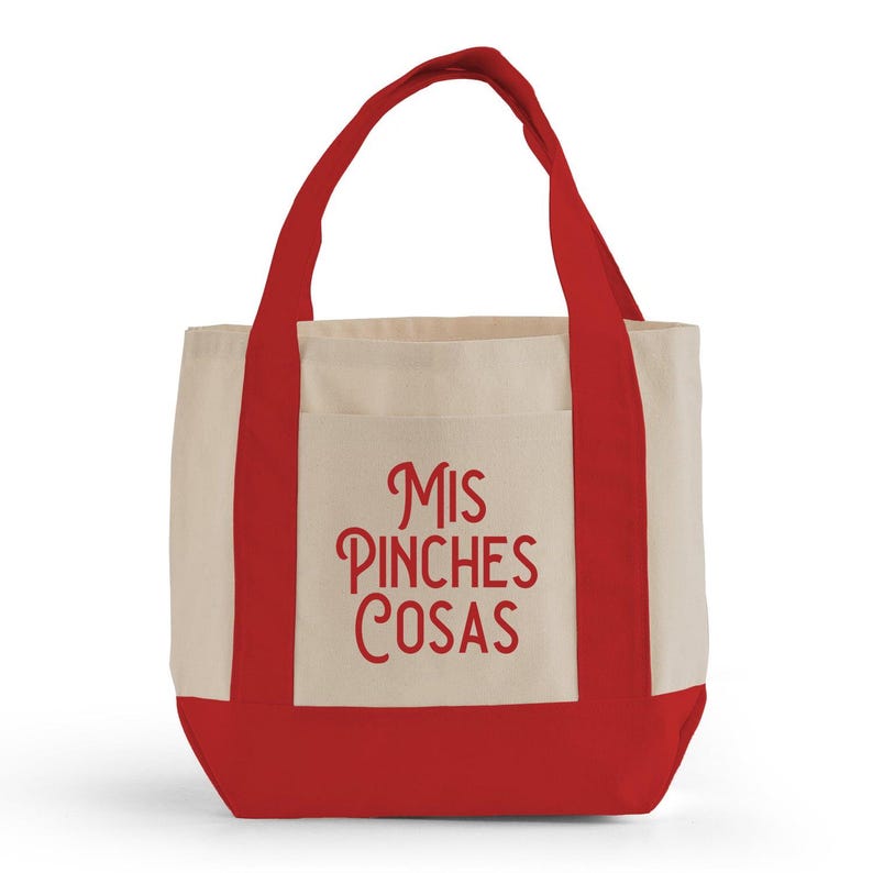 May include: A canvas tote bag with a red base, handles, and trim. The main body is a natural beige color, with a front pocket. The words "MIS PINCHES COSAS" are printed in red script on the front of the bag.