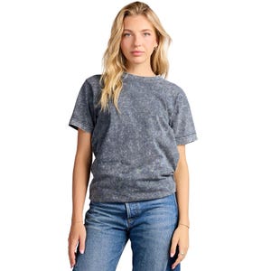 以下が含まれることがあります： ダークチャコールグレーの半袖クルーネックTシャツで、まだら模様のアシッドウォッシュ加工が施されています。袖口と襟元はリブ編みで、標準的なフィット感のシルエットです。ボトムスには、ボタンフライ仕様のクラシックなブルーデニムジーンズを合わせています。