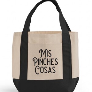 May include: A beige tote bag with black accents and handles. The bag features a front pocket and the phrase "Mis Pinches Cosas" in black script. The bottom of the bag is black.