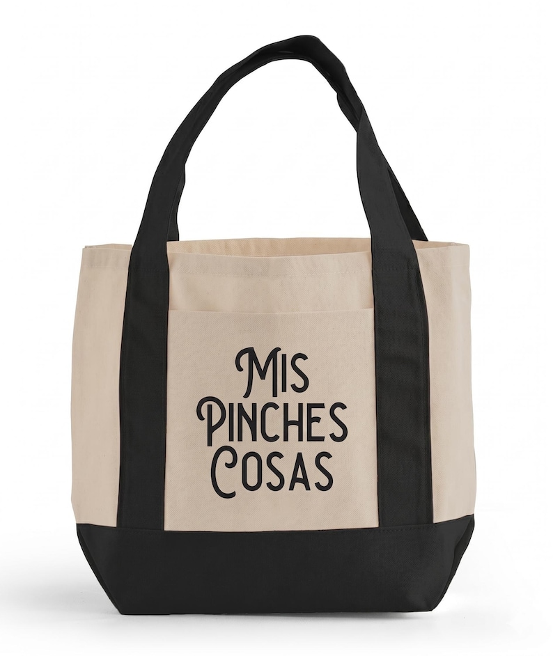 May include: A beige tote bag with black accents and handles. The bag features a front pocket and the phrase "Mis Pinches Cosas" in black script. The bottom of the bag is black.