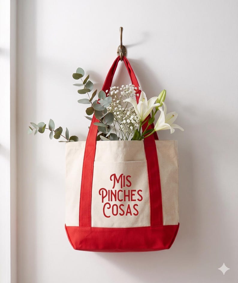 May include: A canvas tote bag with red accents and the text "Mis Pinches Cosas" in red script. The bag is filled with white lilies, baby's breath, and eucalyptus. The bag hangs from a brass hook on a white wall.