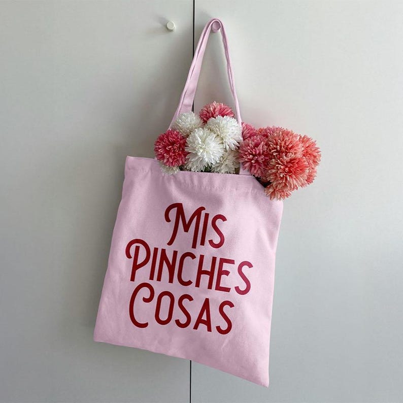 K&ouml;nnte beinhalten: Eine hellrosa Tragetasche mit den Worten "Mis Pinches Cosas" in roter Schrift. Die Tasche ist mit rosa, wei&szlig;en und korallenfarbenen Pomponblumen gef&uuml;llt. Die Tragetasche h&auml;ngt an einer wei&szlig;en T&uuml;r.