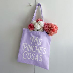 K&ouml;nnte beinhalten: Eine lavendelfarbene Tragetasche mit dem wei&szlig;en Schriftzug "Mis Pinches Cosas". Die Tasche ist mit rosa und wei&szlig;en Pompons gef&uuml;llt. Die Tasche h&auml;ngt an einer wei&szlig;en T&uuml;r.