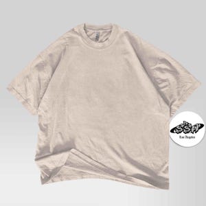 Pu&ograve; includere: Una maglietta oversize beige chiaro con maniche corte e girocollo. La maglietta ha una vestibilit&agrave; ampia. Un logo circolare bianco con la scritta "SSA Los Angeles" si trova nell'angolo in basso a destra.