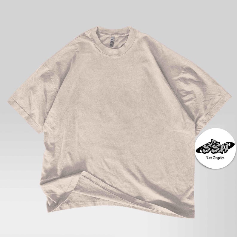 Pu&ograve; includere: Una maglietta oversize beige chiaro con maniche corte e girocollo. La maglietta ha una vestibilit&agrave; ampia. Un logo circolare bianco con la scritta "SSA Los Angeles" si trova nell'angolo in basso a destra.