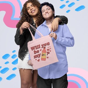 Op de afbeelding: Een lichtroze draagtas met de tekst "Will you be my" en een flesafbeelding met hartjes. De tas wordt door twee personen vastgehouden. E&eacute;n persoon draagt een zwart jasje en een witte rok, de ander een blauw overhemd en een zwarte broek.