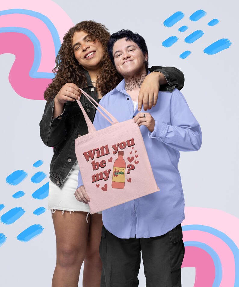 Op de afbeelding: Een lichtroze draagtas met de tekst "Will you be my" en een flesafbeelding met hartjes. De tas wordt door twee personen vastgehouden. E&eacute;n persoon draagt een zwart jasje en een witte rok, de ander een blauw overhemd en een zwarte broek.