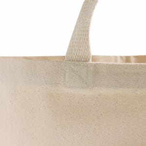 Op de afbeelding: Detail van een natuurlijke canvas draagtas. De tas heeft een stevige, geweven handgreep die aan de bovenrand is bevestigd. Het canvas materiaal heeft een licht getextureerde uitstraling, met een neutrale, gebroken witte kleur. De tas is ontworpen om items te dragen.