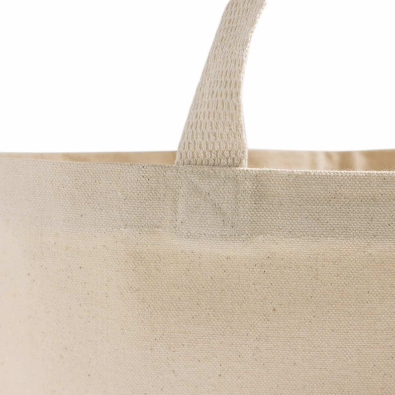 Op de afbeelding: Detail van een natuurlijke canvas draagtas. De tas heeft een stevige, geweven handgreep die aan de bovenrand is bevestigd. Het canvas materiaal heeft een licht getextureerde uitstraling, met een neutrale, gebroken witte kleur. De tas is ontworpen om items te dragen.