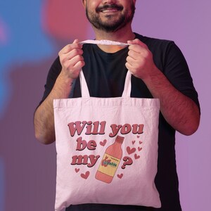 Op de afbeelding: Een witte draagtas met de tekst "Will you be my Valentine?" in roze. De tas heeft een cartoon wijnfles en hartafbeeldingen. De tas wordt vastgehouden door een persoon die een zwart shirt en een spijkerbroek draagt.