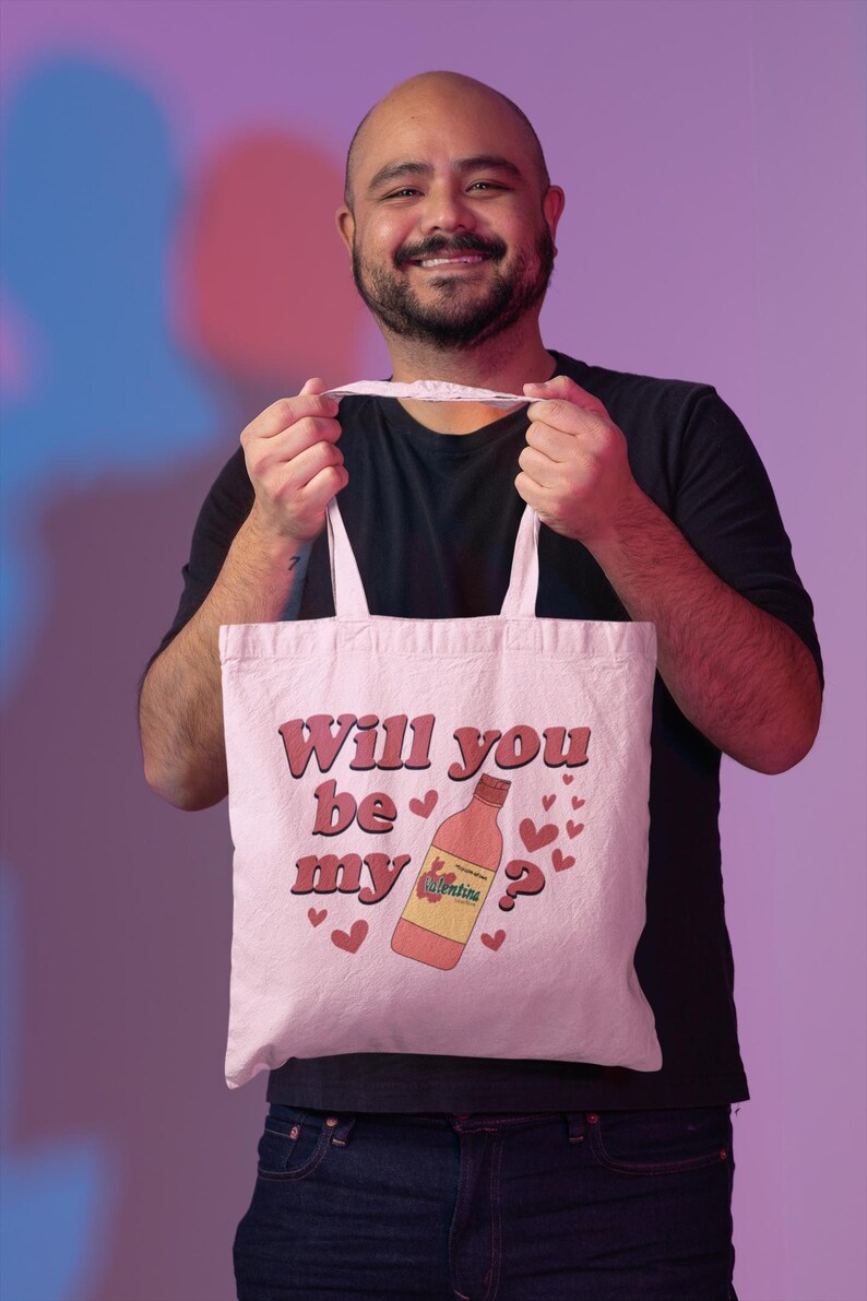 Op de afbeelding: Een witte draagtas met de tekst "Will you be my Valentine?" in roze. De tas heeft een cartoon wijnfles en hartafbeeldingen. De tas wordt vastgehouden door een persoon die een zwart shirt en een spijkerbroek draagt.