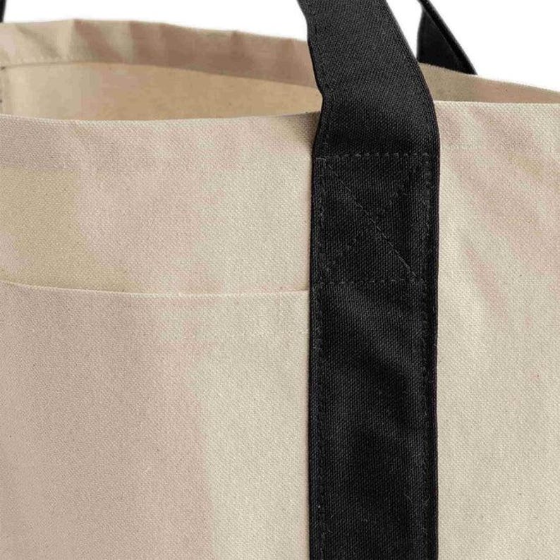 K&ouml;nnte beinhalten: Nahaufnahme einer beigefarbenen Canvas-Tragetasche mit schwarzem Griff. Die Tasche hat ein schlichtes Design mit einer Tasche auf der Vorderseite. Der schwarze Griff ist f&uuml;r die Haltbarkeit an der Tasche befestigt. Die Tasche besteht aus robustem Canvas-Material.