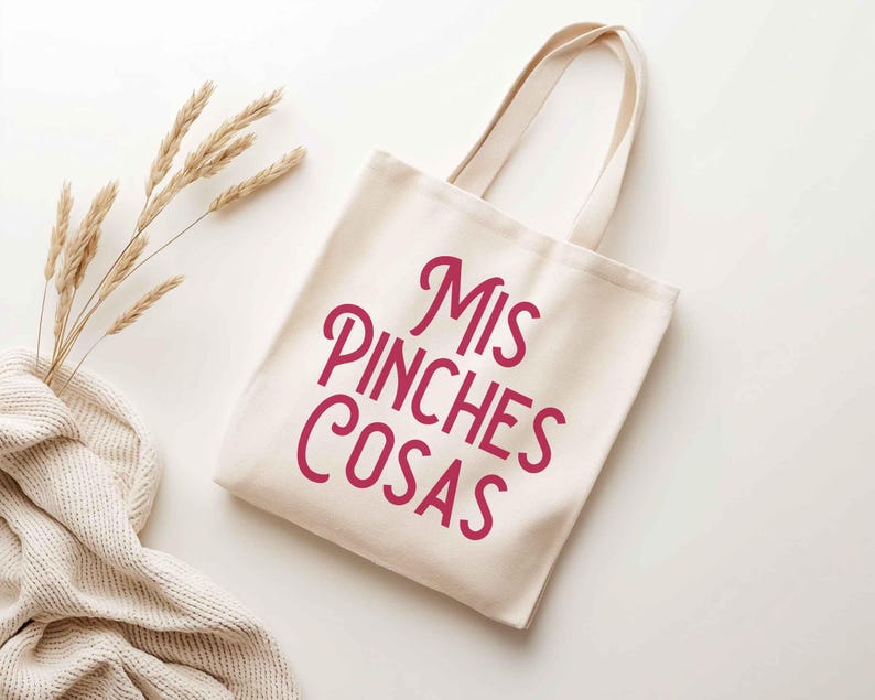 K&ouml;nnte beinhalten: Eine cremefarbene Tragetasche mit dem Text "Mis Pinches Cosas" in einer kr&auml;ftigen, kursiven, magenta Schrift. Die Tasche hat zwei Griffe und ist aus einem robusten Stoff gefertigt. Der Hintergrund ist wei&szlig;, mit einem Zweig aus getrocknetem Weizen und einer cremefarbenen Strickdecke.