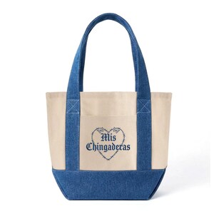 K&ouml;nnte beinhalten: Cremefarbene Tragetasche mit blauen Jeans-Akzenten. Die Tasche hat eine Vordertasche und blaue Jeans-Griffe. Der Schriftzug "Mis Chingaderas" ist in blauer Schrift in einem herzf&ouml;rmigen Design gestickt. Ein stilvolles und praktisches Accessoire.