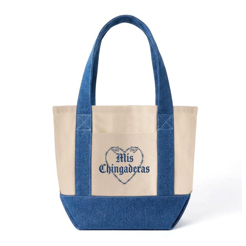 K&ouml;nnte beinhalten: Cremefarbene Tragetasche mit blauen Jeans-Akzenten. Die Tasche hat eine Vordertasche und blaue Jeans-Griffe. Der Schriftzug "Mis Chingaderas" ist in blauer Schrift in einem herzf&ouml;rmigen Design gestickt. Ein stilvolles und praktisches Accessoire.