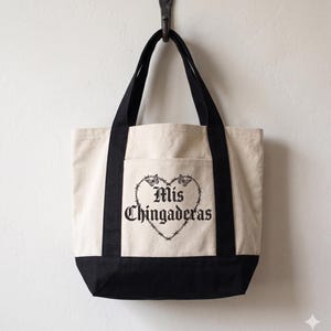 K&ouml;nnte beinhalten: Cremefarbene Tragetasche mit schwarzen Akzenten und Griffen. Die Tasche zeigt ein schwarzes herzf&ouml;rmiges Design mit Stacheldraht und Schmetterlingen sowie den Text "Mis Chingaderas" in einer gotischen Schriftart.