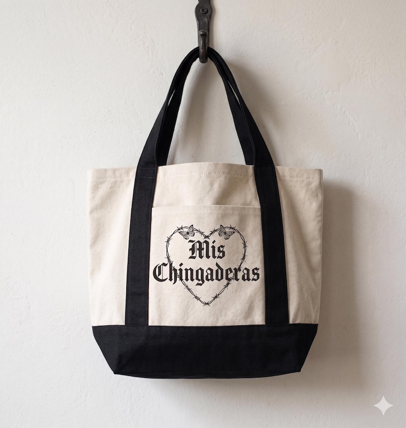 K&ouml;nnte beinhalten: Cremefarbene Tragetasche mit schwarzen Akzenten und Griffen. Die Tasche zeigt ein schwarzes herzf&ouml;rmiges Design mit Stacheldraht und Schmetterlingen sowie den Text "Mis Chingaderas" in einer gotischen Schriftart.