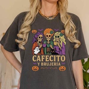 May include: Dark gray t-shirt featuring a Halloween-themed graphic. The design includes three skeleton figures holding mugs, a ghost, a black cat, pumpkins, and the text "CAFECITO Y BRUJER&Iacute;A". The shirt has a relaxed fit.