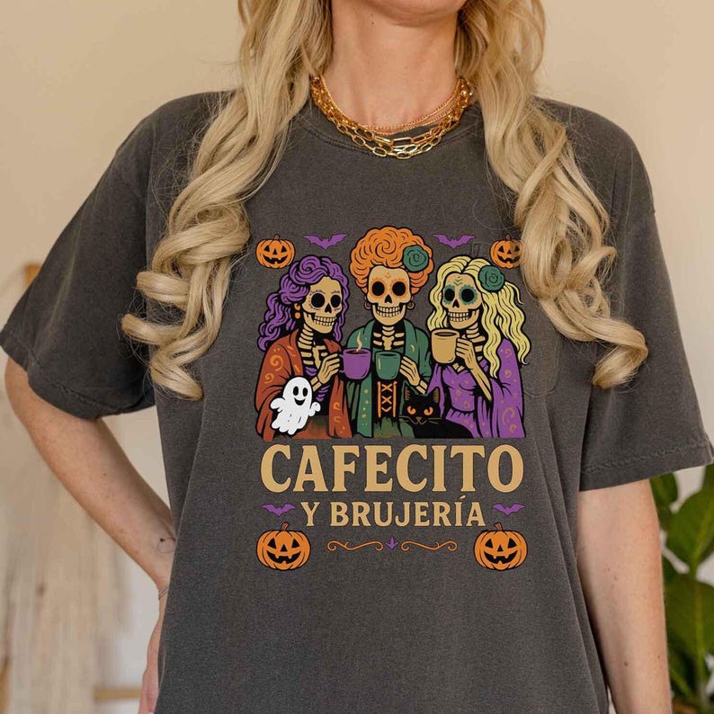 May include: Dark gray t-shirt featuring a Halloween-themed graphic. The design includes three skeleton figures holding mugs, a ghost, a black cat, pumpkins, and the text "CAFECITO Y BRUJER&Iacute;A". The shirt has a relaxed fit.