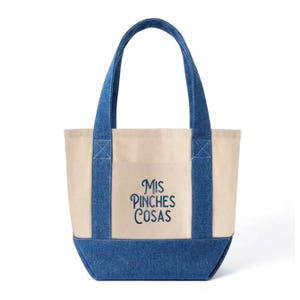 May include: A canvas tote bag with denim blue accents. The bag features a cream-colored body, denim blue handles, and a denim blue bottom. The text "Mis Pinches Cosas" is embroidered in blue on the front. A practical and stylish accessory.
