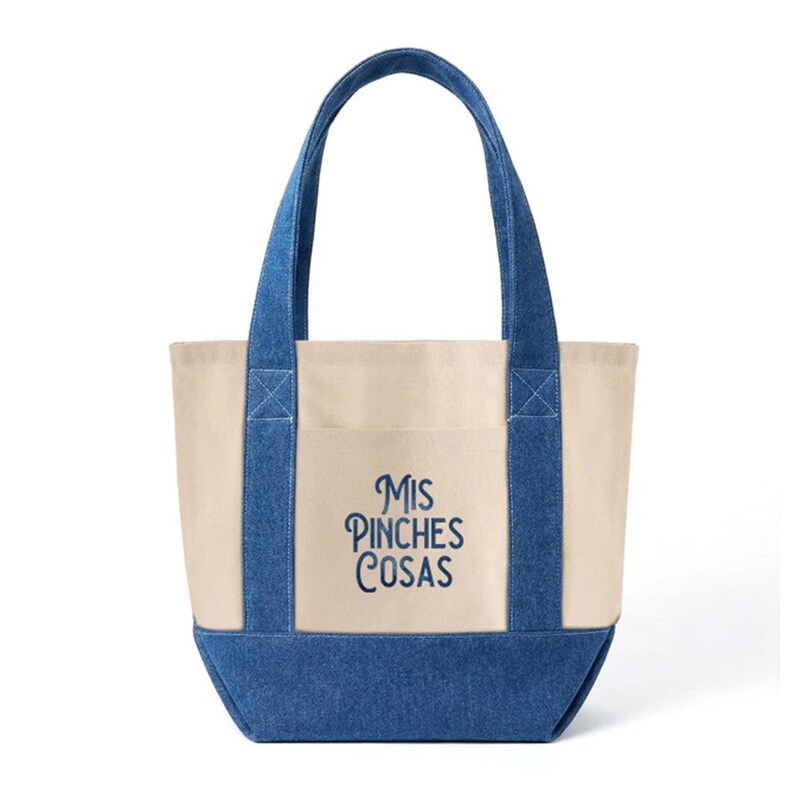 May include: A canvas tote bag with denim blue accents. The bag features a cream-colored body, denim blue handles, and a denim blue bottom. The text "Mis Pinches Cosas" is embroidered in blue on the front. A practical and stylish accessory.
