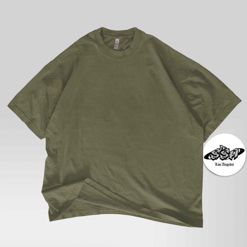Pu&ograve; includere: T-shirt verde oliva a maniche corte, girocollo e vestibilit&agrave; ampia. Un logo bianco e nero con la scritta "Los Angeles" &egrave; in basso a destra.