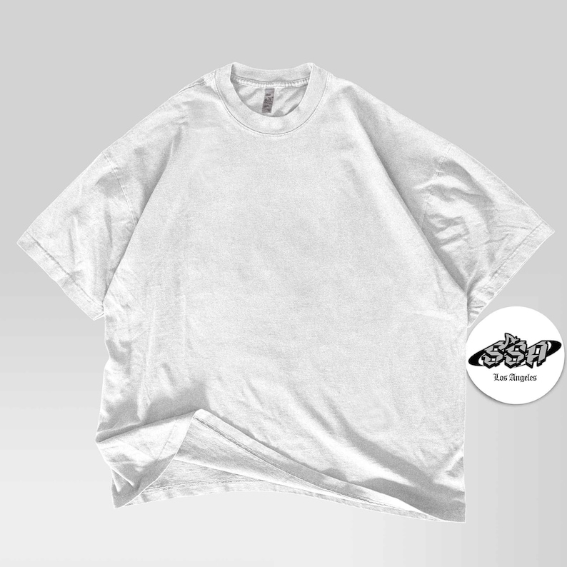 Pu&ograve; includere: Una maglietta bianca semplice, oversize, con girocollo e maniche corte. La maglietta &egrave; realizzata in un tessuto morbido e leggermente strutturato. Un logo circolare con la scritta "SSA Los Angeles" si trova nell'angolo in basso a destra.
