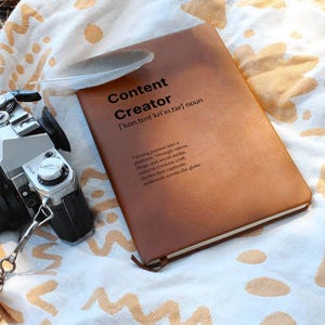 May include: A brown leather-bound journal with the words "Content Creator" and a definition of the term. The journal is lying on a white and tan patterned blanket.