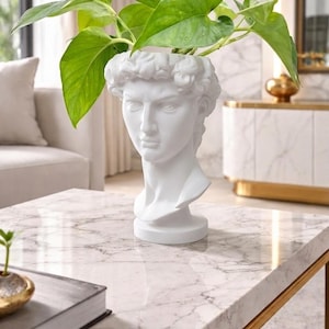 May include: A white bust planter in the shape of a classical male head, holding vibrant green leafy plants. The planter sits on a marble-topped coffee table with a gold-colored metal frame, creating a stylish decor piece.