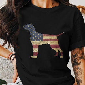 May include: A black t-shirt with a silhouette of a dog in front of an American flag design. The flag is red, white, and blue with a distressed look.