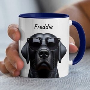 May include: White ceramic mug with a blue rim and handle. The mug features a black Labrador Retriever wearing sunglasses and the name "Freddie" printed on the front.