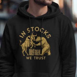 Op de afbeelding: Zwarte hoodie met een gouden afbeelding van een stier en een beer die vechten om een stapel munten. De tekst "In Stocks We Trust" is onder de afbeelding gedrukt.