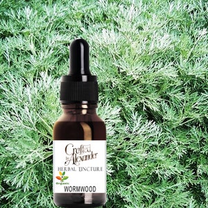 以下が含まれることがあります： ドロップトップ付きの茶色のガラス瓶で、「Crafted by Alexander Herbal Tincture Organic Wormwood」とラベル付けされています。瓶は緑の葉の植物の前にあります。