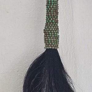 May include: A beaded and fringed horsehair tassel with green and brown beads. The tassel is attached to a leather cord.
