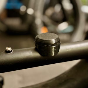 May include: A black, cylindrical component with a textured top and a ridged base, mounted on a black metal tube. A small silver bolt is visible nearby. The background is blurred, suggesting a bicycle frame or similar equipment.