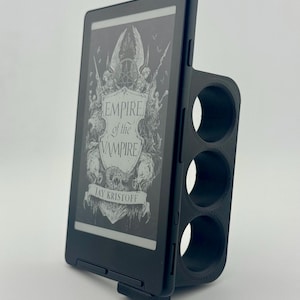 May include: A black e-reader device held in a 3D-printed black plastic grip with three circular finger holes. The screen displays the book cover for Empire of the Vampire by Jay Kristoff, featuring a detailed illustration of a crest and skull.