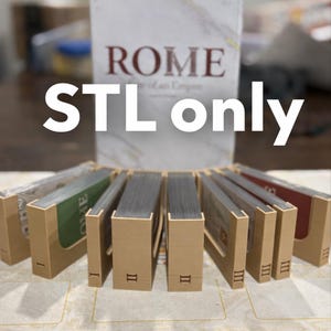 以下が含まれることがあります： 「ROME」というタイトルのボードゲーム。白い文字で「STL only」と書かれています。箱の前には、カードの束が入った複数のタン色のカードホルダーがあります。カードホルダーにはローマ数字が記されています。