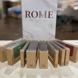 May include: A board game box titled "ROME Fate of an Empire" with a marble-like design. Several card holders are arranged in front, each holding a stack of cards. The card holders are labeled with Roman numerals.